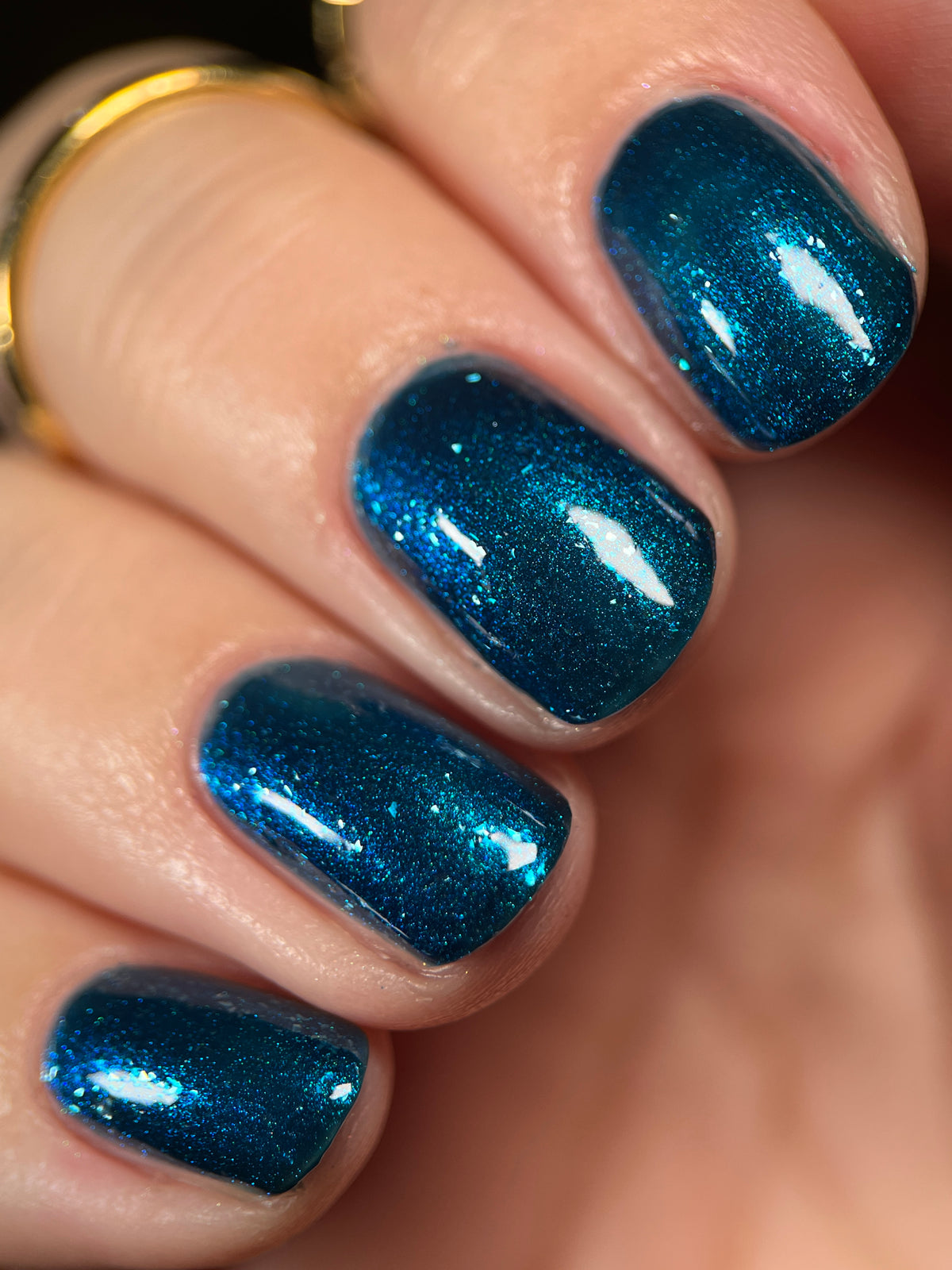 Close-up of fingernails painted with a glittery metallic teal-blue nail polish, shown on a hand with a gold ring visible.