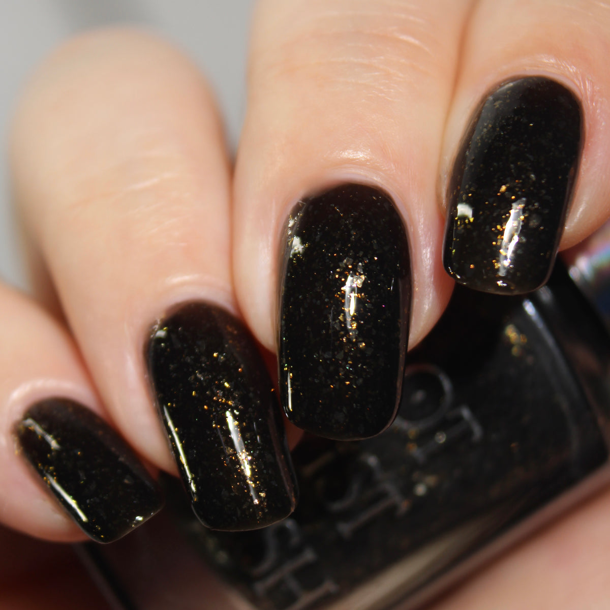 A manicured hand with a black polish with gold flakes holding a bottle of polish.