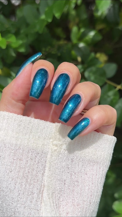A close-up video of a hand with long, almond-shaped nails painted in a shimmering metallic teal-blue nail polish outside in natural sunlight. 